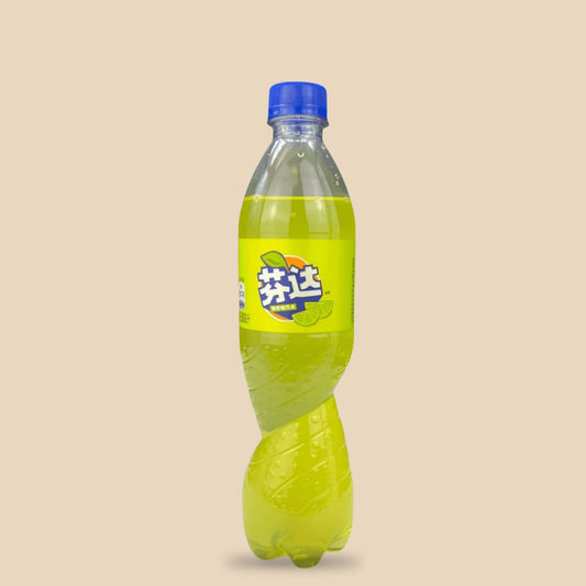 Bouteille verte de Fanta saveur citron vert sur fond beige. Le logo Fanta en caractères chinois blancs et bleus est accompagné des mentions "sans caféine" et "100% arômes naturels". Une illustration de tranche de citron vert juteux est visible sur l'étiquette. La bouteille, ornée de gouttelettes d'eau, évoque une boisson désaltérante, rafraîchissante et fruitée.